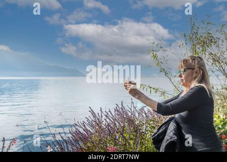 Blonde woman taking a photo with the mobile phone in front of a lake surrounded by mountains. Montreux, Lake Geneva in Switzerland