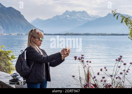 Blonde woman with sunglasses, a jacket and a bag taking a picture of plants on the shore of an alpine lake. Montreux in Geneve lake
