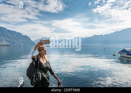 Blonde woman with sunglasses a mask in her arm a selfie in a lake with a boat surrounded by a town and hills with a hard light coming from the sun. Mo Stock Photo