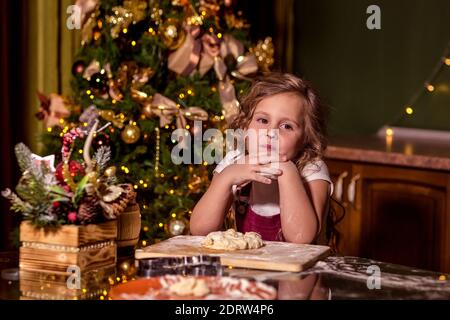 A smiling girl is sitting at a table in the decorated kitchen, with raw dough on the table in front of her. Her nose is smeared with flour. Selective Stock Photo