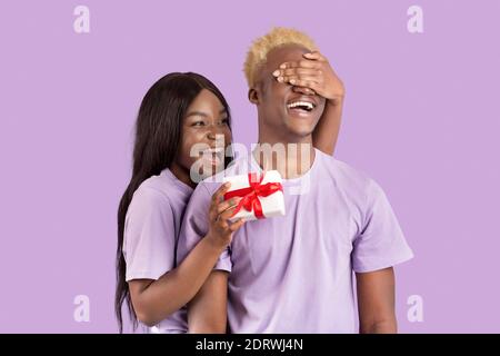 Pretty black woman covering her boyfriend's eyes, surprising him with Valentine's gift on lilac studio background Stock Photo