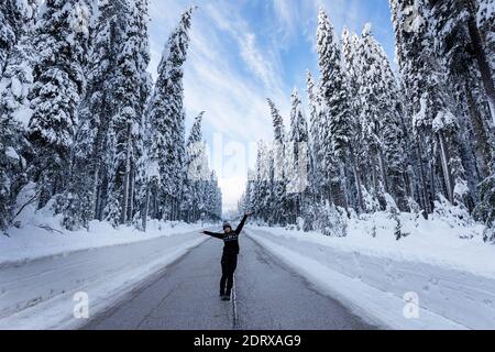 Happy woman raising arms stands in the middle of the road in snow covered forest Stock Photo