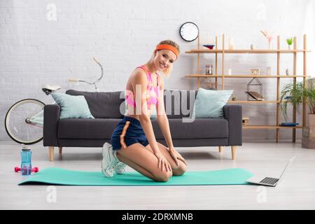 Happy sportswoman looking at camera while sitting on fitness mat near laptop, dumbbells and sports bottle at home Stock Photo