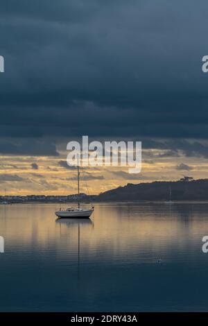 Single Sailing Boat With Reflection In Christchurch Harbour With Hengistbury Head In The Background On A Calm Day At Sunrise With Dark Stormy Clouds Stock Photo