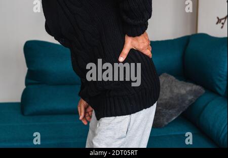 An elderly man touches his back after getting up from the sofa, due to severe pain located in the lower back. Stock Photo