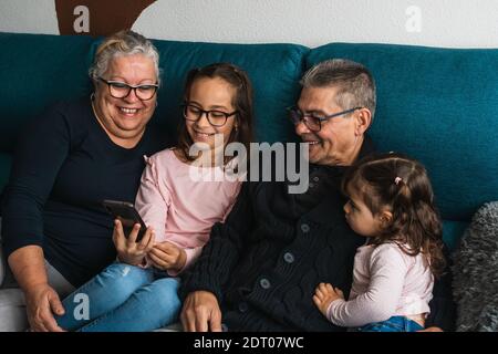Grandfather and grandmother, sitting on the sofa at home with their granddaughters, watching a video on the mobile phone, while having a great time to Stock Photo