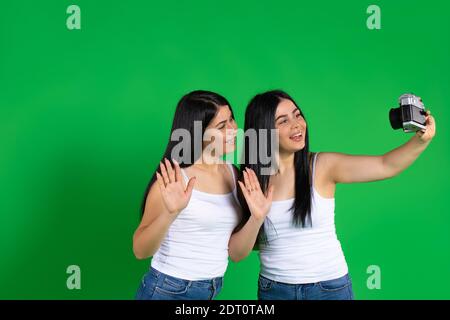 The brunette twins are smiling taking a selfie photo with a vintage camera. Green background. Stock Photo