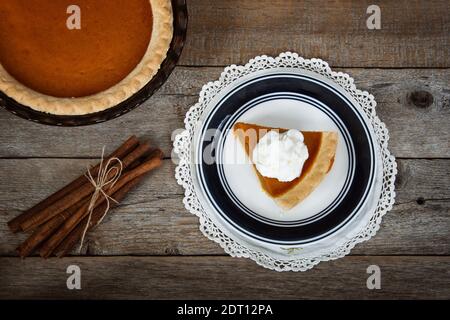 Slice of a pumpkin pie served with whipped cream. Displayed with cinnamon sticks on rustic wooden table. Top view. Stock Photo