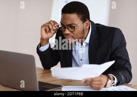 Black guy with glasses accountant holding papers, using laptop Stock Photo
