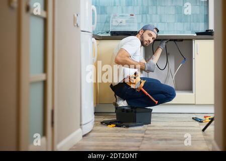 The work is done. Young repairman, professional plumber wearing tool belt looking at camera and showing thumbs up while crouching on the floor, fixing Stock Photo