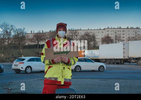 Frankfurt, Germany. December 2020. Young man wearing mask and safety vest holding cardboard arrow direction sign in parking lot. Medium shot. Stock Photo