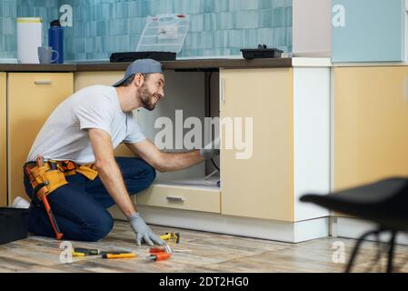 The plumber you can count on. Full length shot of cheerful young plumber wearing tool belt smiling while examining a sink before fixing it in the Stock Photo