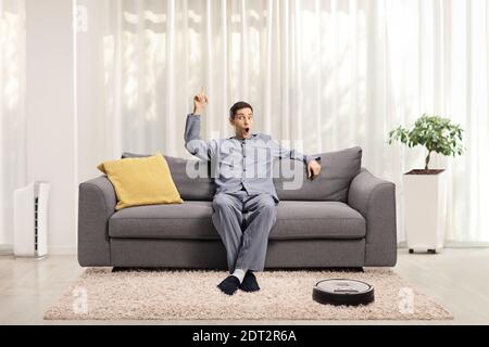 Young excited man in pajamas seated on a sofa at home pointing up and a robotic vacuum cleaner on the floor Stock Photo