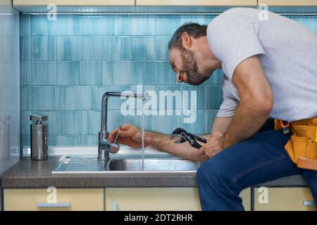 Professional plumber looking concentrated, using pipe wrench while examining and fixing faucet in the kitchen Stock Photo