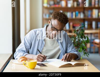 Hardworking black man writing down info from textbook, getting ready for test at cafe Stock Photo