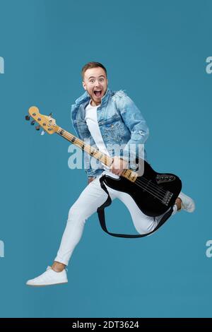 handsome man in denim t-shirt jeans with guitar jumping high isolated on blue background Stock Photo