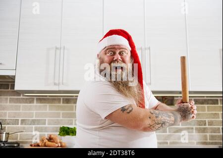An overweigh man in Santa Claus hat with tattooed arms holding a wooden rolling pin on hands like a baseball bate looks at camera thereby showing his Stock Photo