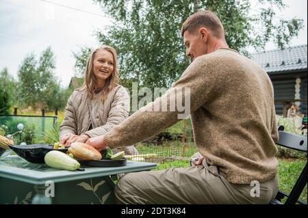 A young couple is preparing for a family barbecue dinner in the courtyard of the house. Cut vegetables on the picnic table. A warm evening in the last days of summer Stock Photo