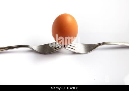 Still life photography. Two forks together create a case for a white egg that balances on them. Egg balanced on forks with white background. Stock Photo
