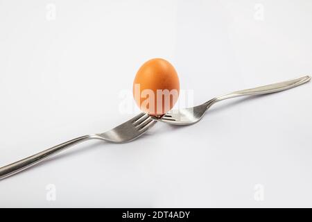 Still life photography. Two forks together create a case for a white egg that balances on them. Egg balanced on forks with white background. Stock Photo