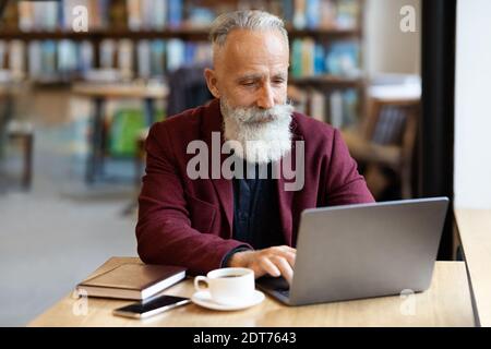 Grey-haired businessman using laptop at cafe, drinking coffee Stock Photo