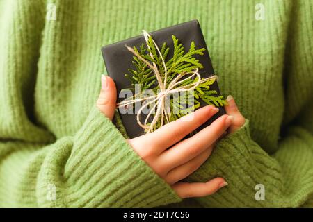 Womans hands holding nice present wrapped in black paper and decorated with green branch, close up Stock Photo
