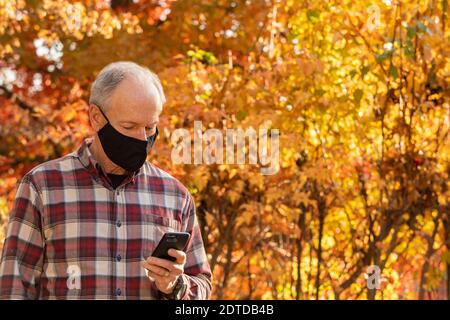 Senior man wearing Covid protective mask using smartphone outdoors in fall Stock Photo