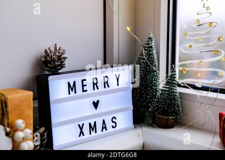 Lightbox with text MERRY XMAS christmas on windowsill. New year decorations, garland lights and christmas trees. Window painted with snow spray. Hygge Stock Photo