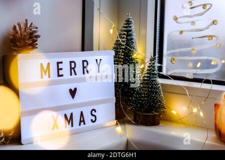 Lightbox with text MERRY XMAS christmas on windowsill. New year decorations, garland lights and christmas trees. Window painted with snow spray. Hygge Stock Photo