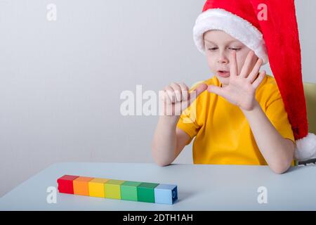boy counting on his fingers sitting at a table in a santa hat. on the table colorful shapes blocks in a line Stock Photo