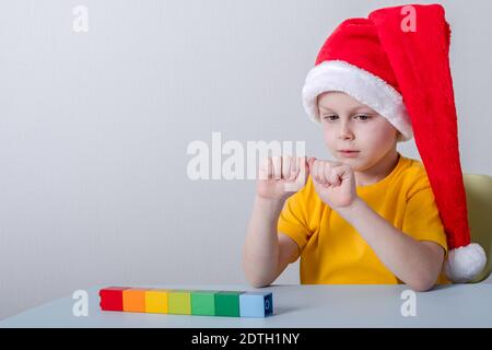 boy counting on his fingers sitting at a table in a santa hat. on the table colorful shapes blocks in a line Stock Photo