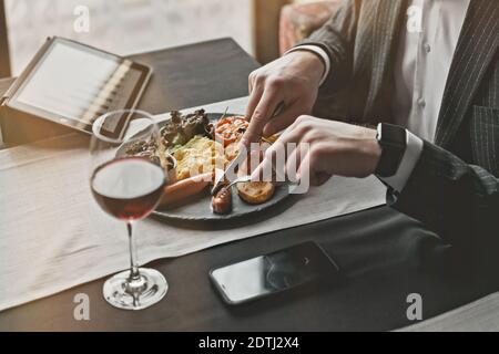 A young and attractive man uses his tablet while eating Traditional English breakfast. Sausage, beans, eggs, bread and salad in an indoor restaurant. Stock Photo