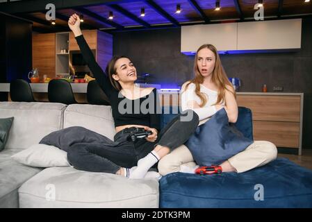Attractive cheerful young female friends sitting on comfortable couch in the modern apartment and enjoying video games. One girl feels happy winning, Stock Photo