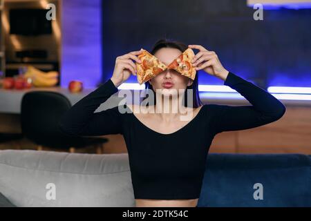 Excited happy young woman in kitchen playing with pizza slices, smiling and covered her eyes. Carefree girl very playful. Pizza delivery online. Stock Photo