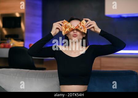 Carefree playful young woman in kitchen playing with pizza slices, smiling and covered her eyes. Pizza delivery online. Unhealthy eating concept. Stock Photo