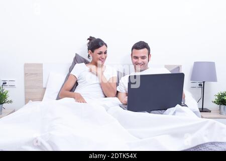 Happy laughing relaxed young couple using laptop in bed at home. Intermet and work at home concept Stock Photo