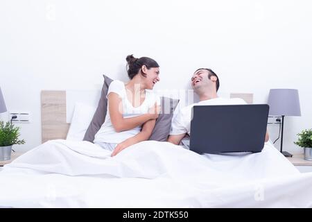 Happy laughing relaxed young couple using laptop in bed at home. Intermet and work at home concept Stock Photo