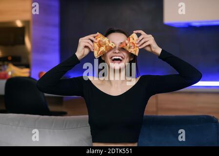 Excited happy young woman in kitchen playing with pizza slices, smiling and covered her eyes. Carefree girl very playful. Pizza delivery online. Stock Photo