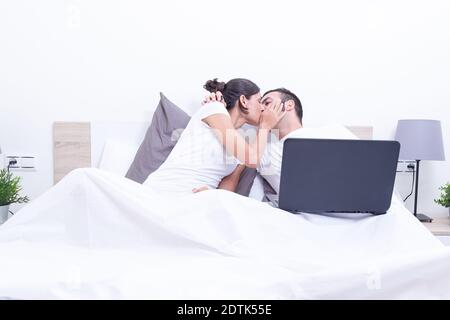 Happy  young couple kissing in bed using laptop at home. Intermet, work at home and love concept Stock Photo