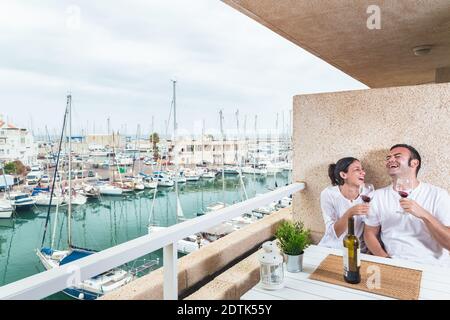 Happy  young couple smiling, drinking red wine on the terrace at the marina. Couple relationship and lifestyle concept. Stock Photo
