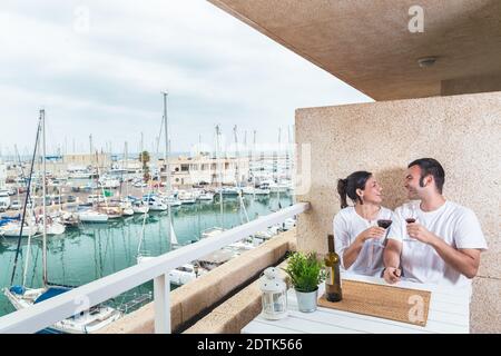 Happy  young couple smiling, drinking red wine on the terrace at the marina. Couple relationship and lifestyle concept. Stock Photo