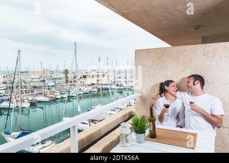 Happy  young couple smiling, drinking red wine on the terrace at the marina. Couple relationship and lifestyle concept. Stock Photo