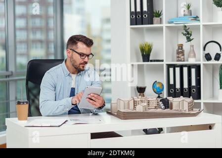 An experienced male architect in office inspects the project of a residential complex and makes calculations using a tablet PC. Stock Photo