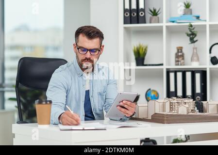 An experienced male architect in office inspects the project of a residential complex and makes calculations using a tablet PC. Stock Photo