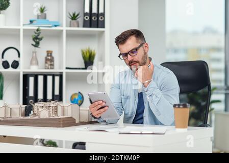 An experienced male architect in office inspects the project of a residential complex and makes calculations using a tablet PC. Stock Photo