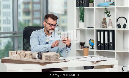 An experienced male architect in office inspects the project of a residential complex and makes calculations using a tablet PC. Stock Photo