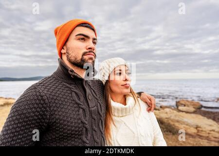 Handsome guy embracing his girlfriend outdoor on a sea resort during winter vacation watching horizon at sunset wearing sweater and wool cap for cold Stock Photo