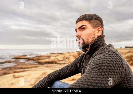 Handsome Caucasian serious young man sitting on sea rocks at sea resort watching horizon wearing sweater - Stylish guy with pensive glance with cloudy Stock Photo