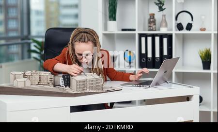 Female architect with dreadlocks makes measurements from mock-up of new buildings in architectural bureau. Stock Photo