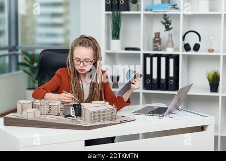 Female architect with dreadlocks makes measurements from mock-up of new buildings in architectural bureau. Stock Photo